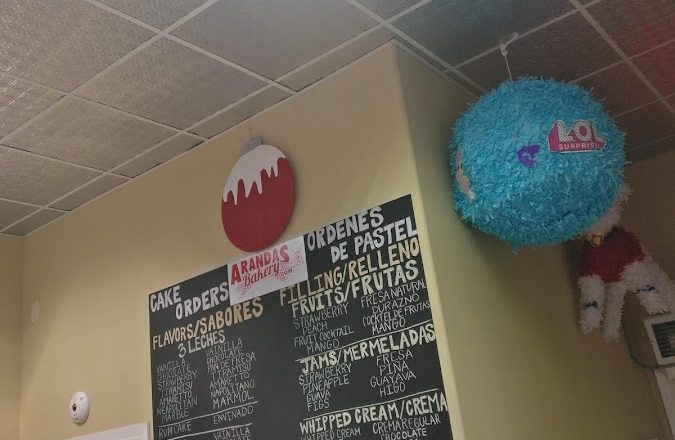 Interior with chalkboard cake order menu listing flavors, fillings, and toppings with the Arandas Bakery logo and colorful piñatas hanging from the ceiling at Arandas Bakery