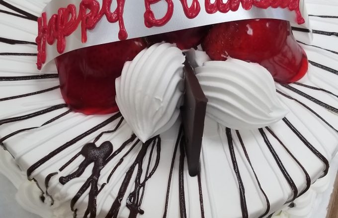 Round white birthday cake with chocolate drizzle, strawberries, whipped cream rosettes, chocolate shards, and a Happy Birthday banner in a box at Arandas Bakery