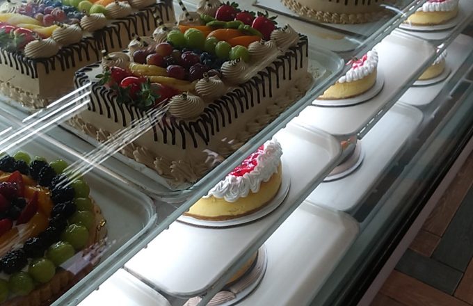 Glass display case filled with decorated fruit cakes, round cakes with chocolate drizzle, fruit tarts, and small pastries at Arandas Bakery