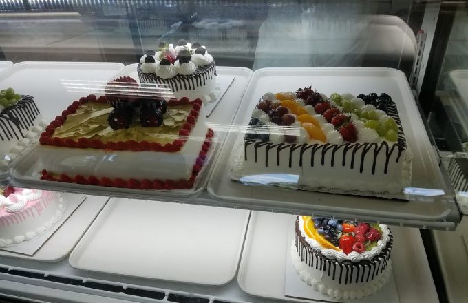 Refrigerated display case with rectangular and round fruit cakes topped with berries, grapes, and chocolate drizzle at Arandas Bakery