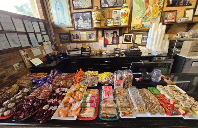 Counter display of colorful packaged Mexican candies and dulces with family photos and artwork on the wall at La Mexicana