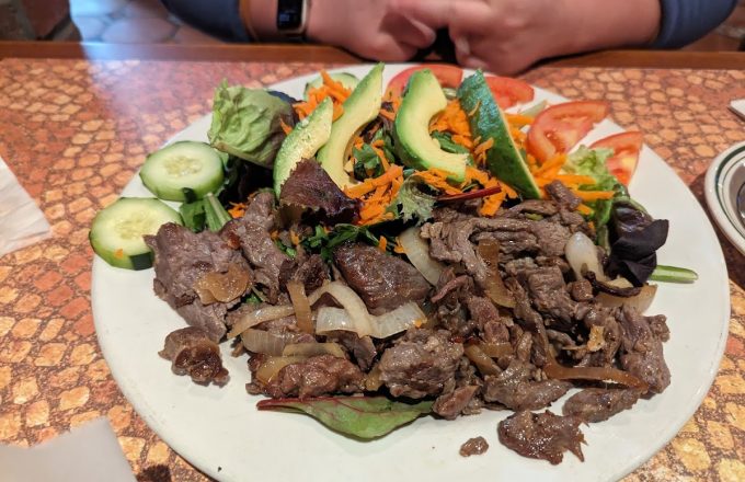 Grilled beef with sautéed onions, sliced avocado, cucumber, shredded carrots, and tomato on a white plate at La Mexicana