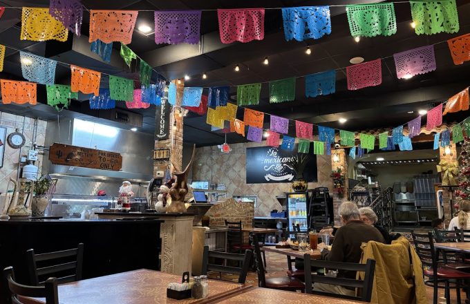 Dining room with colorful papel picado banners, dark wooden tables, open kitchen, and La Mexicana sign on the wall at La Mexicana