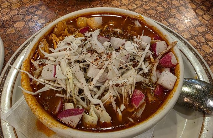 Bowl of red pozole topped with shredded cabbage, diced radish, and oregano on a mosaic table at La Mexicana