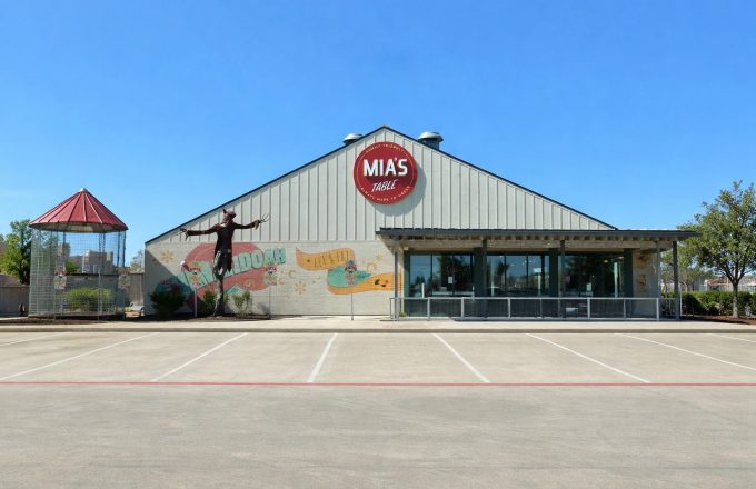 Exterior of Mia's Table Shenandoah location with colorful Let's Eat mural, round Mia's Table sign, and covered patio entrance