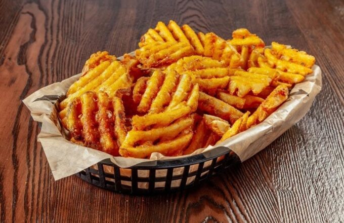 Basket of golden waffle-cut fries in a black plastic basket on a dark wood table