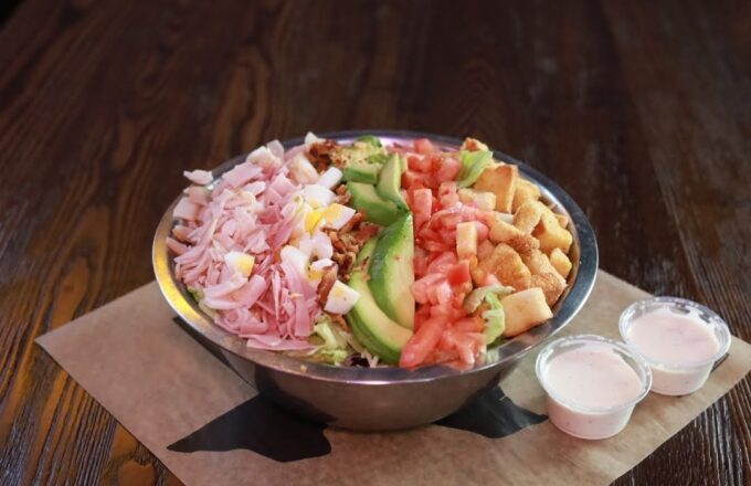 Cobb salad in a stainless steel bowl with ham, avocado, tomato, egg, and croutons, with two cups of dressing on the side