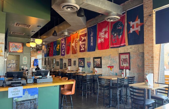 Texadelphia Garden Oaks interior — bar seating with Texas sports flags (Astros, Texans, UH, Aggies, Longhorns) hanging from the ceiling